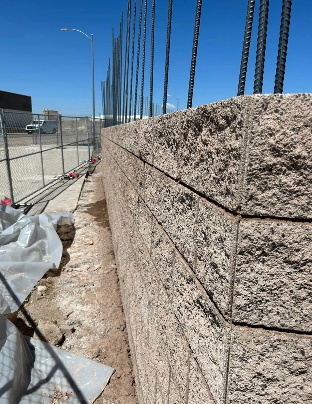 A split-face concrete block wall going up along a chain-link fence, with vertical rebar standing above the top course.